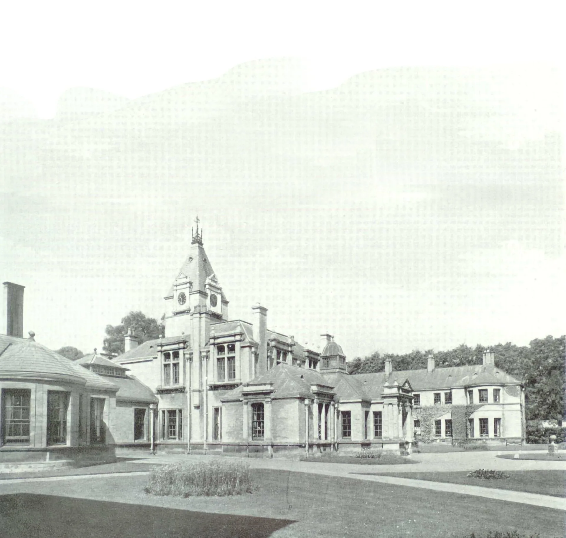 Black and white photo of a large Victorian-era building with a central tower, tall windows, and multiple wings, surrounded by grass, pathways, and trees in the background.